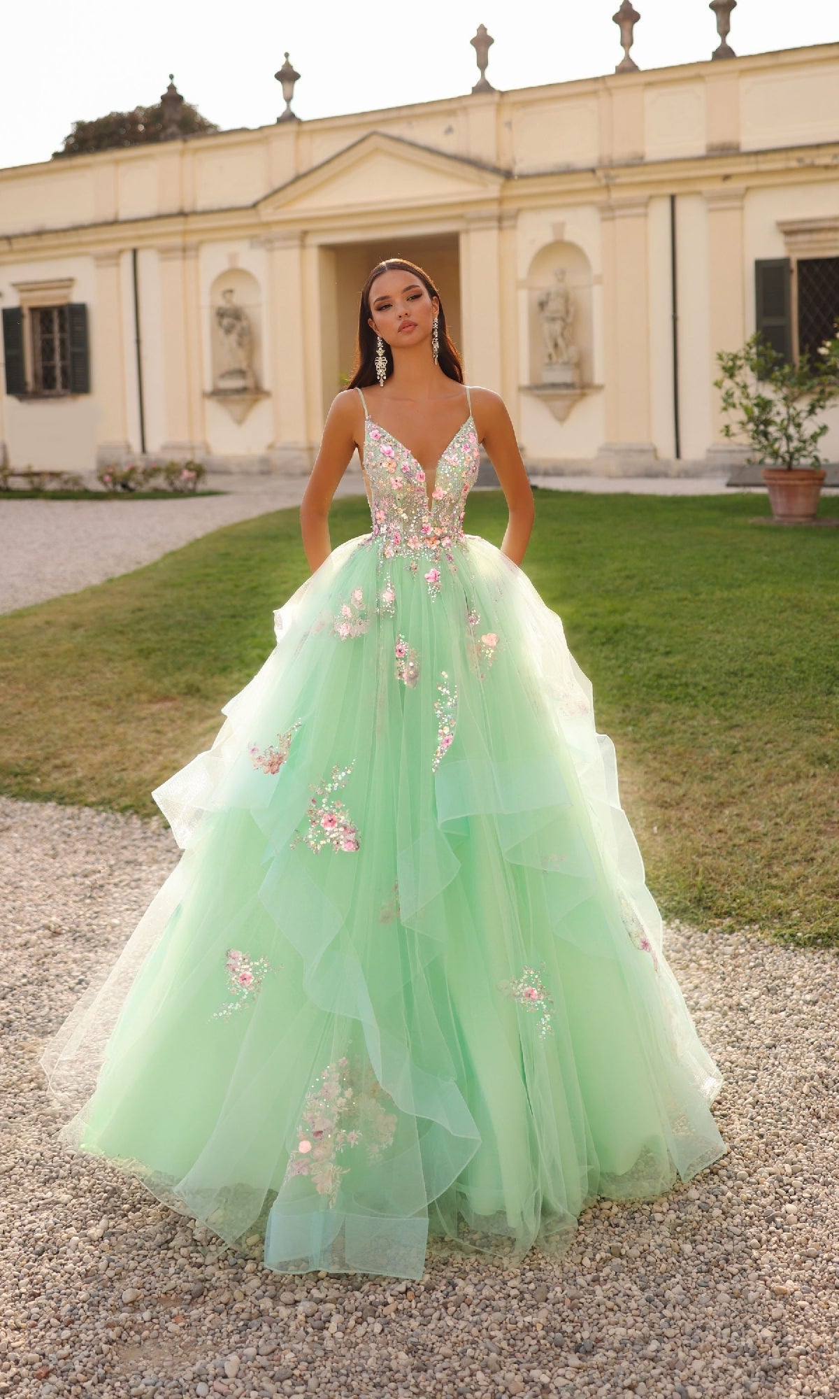 A woman stands outside a classical building wearing the Alyce 61735 long prom dress—a mint green gown with floral embellishments and a layered tulle skirt—looking directly at the camera.
